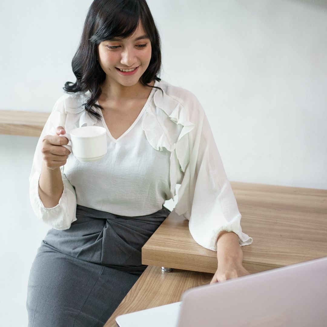 woman in blouse sits at desk with laptop and mug
